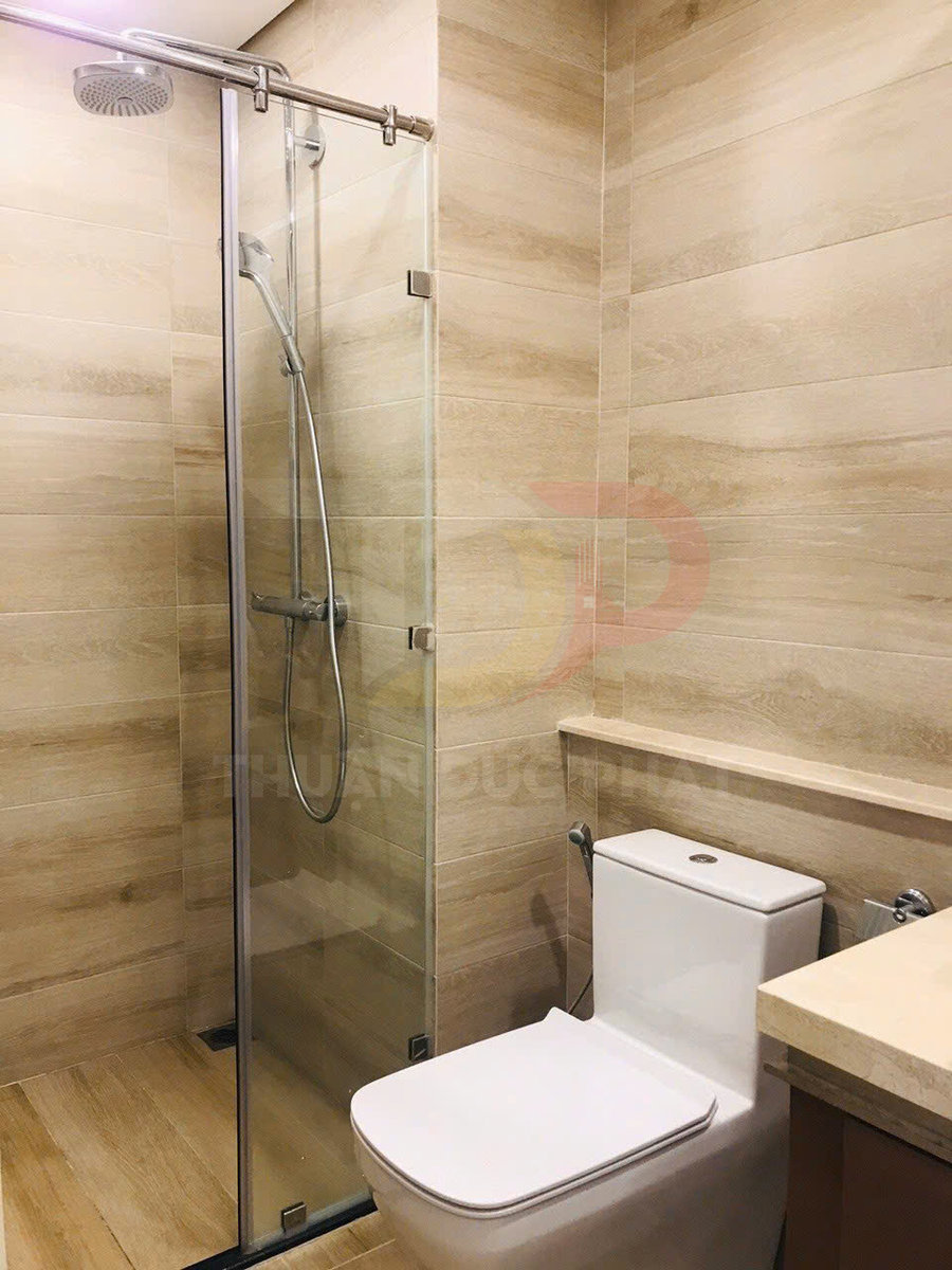 Bồn cầu sứ trắng cạnh buồng tắm kính đứng với vòi sen inox trong phòng tắm ốp gạch giả gỗ