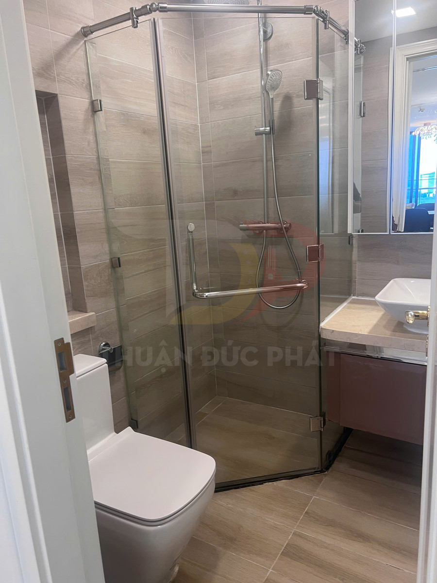 Phòng tắm nhỏ với buồng tắm kính góc, vòi sen tay, bồn cầu sứ trắng và lavabo đặt trên mặt đá