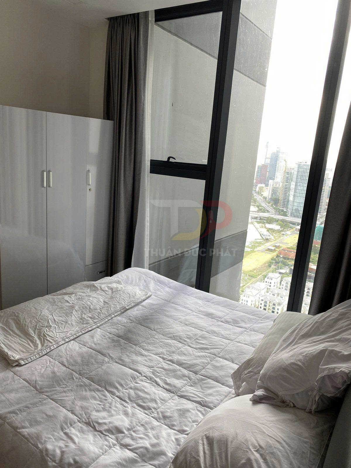 Giường ngủ với chăn ga gối trắng cạnh tủ quần áo và cửa sổ kính lớn nhìn ra thành phố