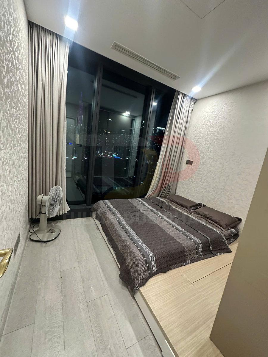 Phòng ngủ nhỏ với giường nệm ga màu nâu cạnh cửa kính lớn nhìn ra thành phố ban đêm