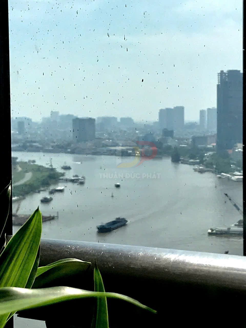 Cửa kính bị bẩn nhìn ra sông và tàu thuyền, phía trước là lan can và chậu cây xanh