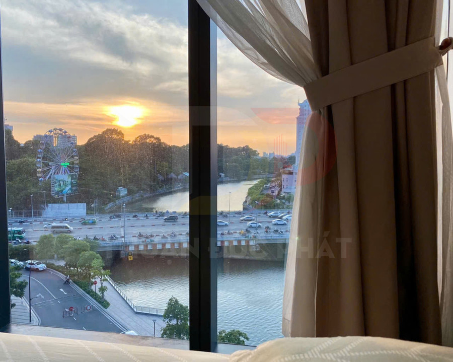 View thành phố nhìn từ cửa sổ phòng ngủ với rèm vải mở ra cảnh sông và đường xe cộ lúc hoàng hôn