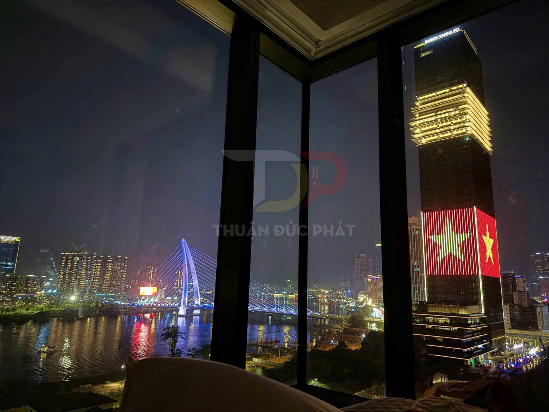 View phòng khách sạn nhìn ra sông Hàn Đà Nẵng, cầu dây văng và tòa nhà cao tầng chiếu cờ Việt Nam ban đêm